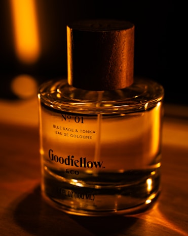 A glass bottle of cologne with a wooden cap is illuminated by warm, ambient lighting, creating a cozy and sophisticated atmosphere. The label on the bottle reads 'Goodfellow & Co' and specifies the scent as 'Blue Sage & Tonka'.
