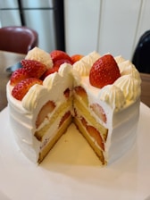 A round cake with white frosting and strawberries adorning the top. The cake is cut to reveal layers of sponge and cream, with whole strawberries embedded within the layers.