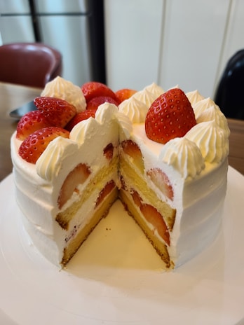 A round cake with white frosting and strawberries adorning the top. The cake is cut to reveal layers of sponge and cream, with whole strawberries embedded within the layers.