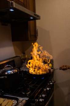 A kitchen stove with a pot and a pan. The pan is positioned on a burner with a large flame rising from it. A hand is holding the pan's handle. The scene is in a dimly lit kitchen with a wooden cabinet and backsplash visible in the background.
