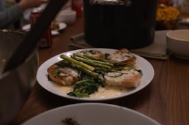 A plate of food consisting of grilled salmon topped with a creamy sauce, accompanied by asparagus and saut&eacute;ed greens, is placed on a wooden dining table. In the background, there are a pot, some bowls, and a blurred soda can, suggesting a meal setting.