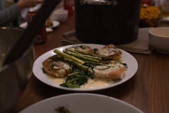 A plate of food consisting of grilled salmon topped with a creamy sauce, accompanied by asparagus and saut&eacute;ed greens, is placed on a wooden dining table. In the background, there are a pot, some bowls, and a blurred soda can, suggesting a meal setting.