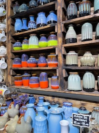 An assortment of colorful ceramic vases and pots are displayed in a wooden shelf. The ceramics come in bright colors such as blue, orange, green, and brown, with various patterns and textures. A small chalkboard sign reads 'Made in Vietnam' with a price range. Additionally, several ceramic animal figures resembling chickens are placed on a lower shelf amidst more blue vases.
