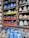 An assortment of colorful ceramic vases and pots are displayed in a wooden shelf. The ceramics come in bright colors such as blue, orange, green, and brown, with various patterns and textures. A small chalkboard sign reads 'Made in Vietnam' with a price range. Additionally, several ceramic animal figures resembling chickens are placed on a lower shelf amidst more blue vases.