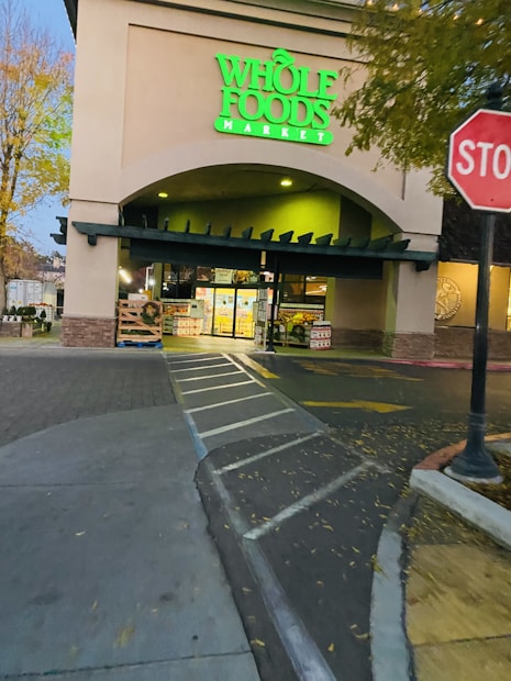 An entrance to a Whole Foods Market with a neon green sign that stands out. The foreground shows a parking area with marked pedestrian lanes and a stop sign. The area is surrounded by trees with yellowing leaves, suggesting an autumn setting. The store displays are visible through the entrance, illuminated by interior lighting.