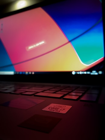 A laptop keyboard is visible with an open screen displaying a blurred multicolored desktop background. The colors are vivid, with prominent shades of blue, orange, and red. An Intel sticker is in focus on the laptop's palm rest area.