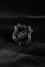 A metallic lapel pin featuring an angular shield shape with the letters 'NHL' prominently displayed in the center. The pin rests against a dark, textured fabric background that enhances its shiny, reflective surface.