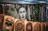A tapestry with a black and white portrait of a woman surrounded by floral patterns hangs in the background. In the foreground, colorful handmade designs including a skull and circular patterns are displayed on canvas. The setup appears to be in an outdoor market setting, with trees visible in the background.