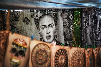 A tapestry with a black and white portrait of a woman surrounded by floral patterns hangs in the background. In the foreground, colorful handmade designs including a skull and circular patterns are displayed on canvas. The setup appears to be in an outdoor market setting, with trees visible in the background.