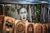 A tapestry with a black and white portrait of a woman surrounded by floral patterns hangs in the background. In the foreground, colorful handmade designs including a skull and circular patterns are displayed on canvas. The setup appears to be in an outdoor market setting, with trees visible in the background.