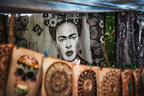 A tapestry with a black and white portrait of a woman surrounded by floral patterns hangs in the background. In the foreground, colorful handmade designs including a skull and circular patterns are displayed on canvas. The setup appears to be in an outdoor market setting, with trees visible in the background.