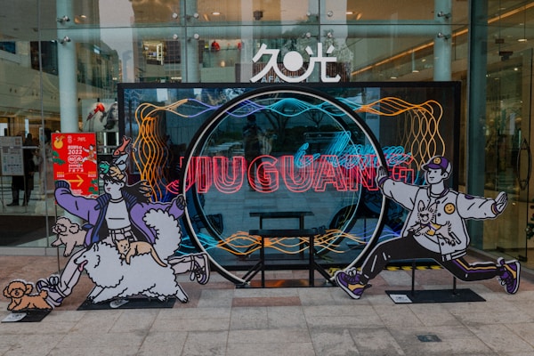 Colorful artistic displays are set in front of a glass storefront. Two large cut-out illustrations of cartoon-like characters are positioned on either side of a circular neon installation in the center. The characters are dynamically posed, depicting a person with animals, such as a small dog and a cat, harmoniously integrated into the design. Vibrant neon lights spell out text, and an advertisement poster is visible on the left.