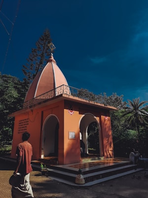 A small temple-like structure with an orange facade and a dome on top. The scene is set against a clear blue sky, surrounded by trees including a palm and a pine. Two arches form the entrance, and there is text written on the side of the building. A person in traditional attire, possibly holding a vessel, is walking near the entrance.