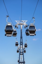 Multiple cable cars are suspended on cables against a bright blue sky, suggesting a high-altitude transportation system in motion. The cable cars are structured in a line along the cables, indicating organized movement.