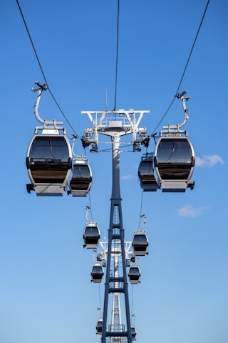Multiple cable cars are suspended on cables against a bright blue sky, suggesting a high-altitude transportation system in motion. The cable cars are structured in a line along the cables, indicating organized movement.