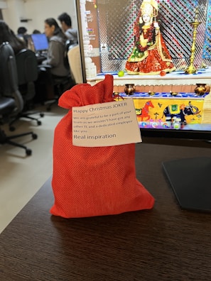 A desk scene with a red fabric gift pouch in the foreground. The pouch has a message attached that reads 'Happy Christmas JOKER... Real inspiration.' Behind the pouch is a computer monitor displaying an image of a deity in traditional attire. Several people are seated at computers in the background, suggesting an office environment.