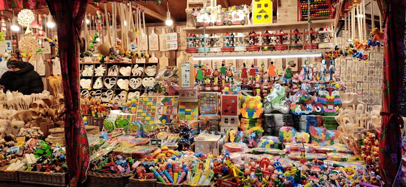 A festive market stall is filled with a variety of colorful toys and handcrafted items. Shelves and tables are crowded with wooden puzzles, decorative figures, heart-shaped ornaments, and stacking toys. Baskets on the lower sections contain more toys, and the overall scene is vibrant and lively.