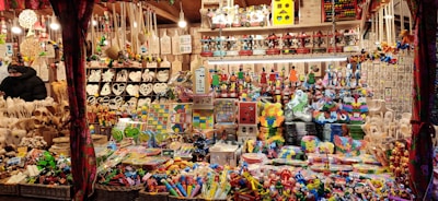 A festive market stall is filled with a variety of colorful toys and handcrafted items. Shelves and tables are crowded with wooden puzzles, decorative figures, heart-shaped ornaments, and stacking toys. Baskets on the lower sections contain more toys, and the overall scene is vibrant and lively.
