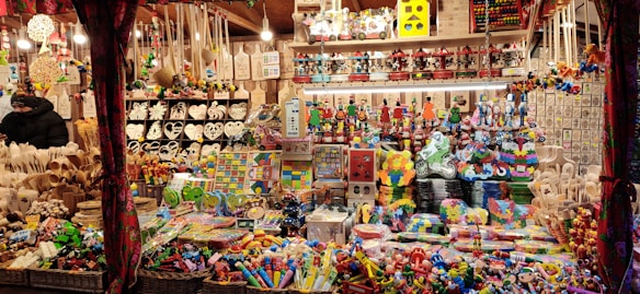 A festive market stall is filled with a variety of colorful toys and handcrafted items. Shelves and tables are crowded with wooden puzzles, decorative figures, heart-shaped ornaments, and stacking toys. Baskets on the lower sections contain more toys, and the overall scene is vibrant and lively.