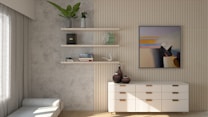 A modern interior space featuring a textured wall with two minimalist floating shelves. The shelves display a variety of decor items, including books, plants, and a globe. A contemporary abstract painting hangs on the wall above a sleek white dresser with wooden handles. On the dresser are two vases that complement the neutral color palette. A soft, light-colored bench is positioned near a window with sheer curtains allowing natural light to illuminate the room.