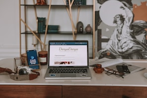 An aesthetically arranged workspace featuring a laptop displaying a webpage, a smartphone, a camera, a pair of glasses, and a few decorative items like candles and pottery. The background includes a wall with a shelf holding books and artful objects, alongside a large black-and-white artwork.
