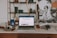 An aesthetically arranged workspace featuring a laptop displaying a webpage, a smartphone, a camera, a pair of glasses, and a few decorative items like candles and pottery. The background includes a wall with a shelf holding books and artful objects, alongside a large black-and-white artwork.