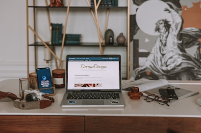 An aesthetically arranged workspace featuring a laptop displaying a webpage, a smartphone, a camera, a pair of glasses, and a few decorative items like candles and pottery. The background includes a wall with a shelf holding books and artful objects, alongside a large black-and-white artwork.