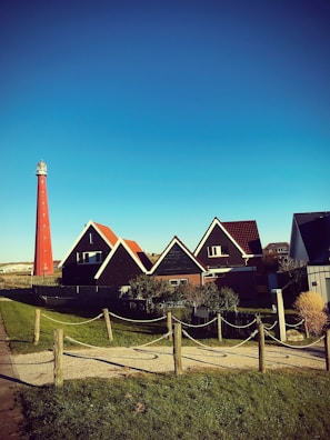 A picturesque landscape featuring a bright red lighthouse standing tall against a vibrant blue sky. In the foreground, charming houses with distinct red and brown roofs are nestled along a well-kept path that is bordered by neatly trimmed green grass. A rustic fence with light ropes adds to the quaint, serene atmosphere of the scene.