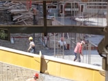 A construction site with workers wearing helmets engaged in labor. Scattered materials and scaffolding surround the area, with notable metal rods protruding from the ground. An exit sign is visible in the foreground.