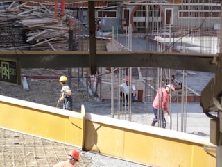 A construction site with workers wearing helmets engaged in labor. Scattered materials and scaffolding surround the area, with notable metal rods protruding from the ground. An exit sign is visible in the foreground.