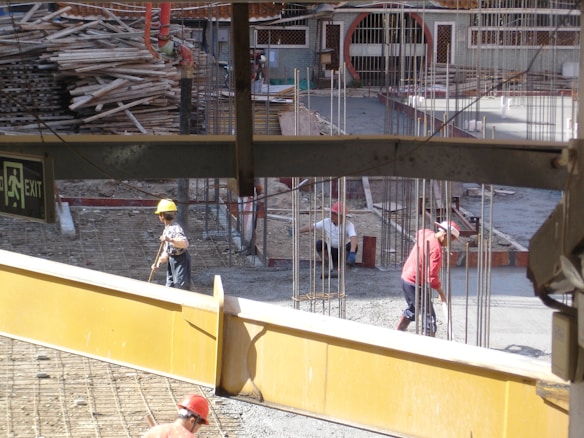 A construction site with workers wearing helmets engaged in labor. Scattered materials and scaffolding surround the area, with notable metal rods protruding from the ground. An exit sign is visible in the foreground.