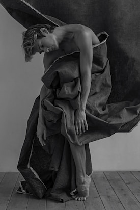 A person is wrapped in a large, flowing fabric that drapes around their body. The artistic pose highlights the figure's physique and grace. The setting is minimalist, with a wooden floor and a plain background, creating a serene and introspective atmosphere.