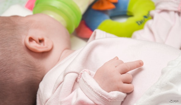 A baby lying on a soft surface with a bottle nearby, dressed in light pink clothing. The baby's hand is prominently visible, and a colorful plush or toy ring with green, blue, and orange hues is in the background.