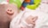 A baby lying on a soft surface with a bottle nearby, dressed in light pink clothing. The baby's hand is prominently visible, and a colorful plush or toy ring with green, blue, and orange hues is in the background.