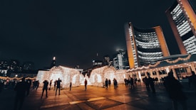 A night scene in a city plaza with an illuminated installation made from numerous small lights in the shape of arches. Tall buildings with lit windows surround the area, and a crowd of people is visible, walking and observing the lights.