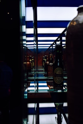 A collection of vintage bottles is displayed on mirrored shelves, creating a reflective effect. The background features horizontal blue and white stripes, adding a modern touch. The image has a mix of dim and illuminated lighting creating a contrast.