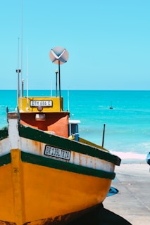 A vibrant yellow and green fishing boat is positioned on a concrete surface near a bright, turquoise-colored sea. The boat features visible registration plates and tall antennas. The weather appears sunny, with a clear blue sky. The calm ocean provides a serene background.