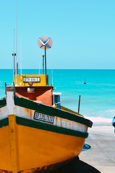 A vibrant yellow and green fishing boat is positioned on a concrete surface near a bright, turquoise-colored sea. The boat features visible registration plates and tall antennas. The weather appears sunny, with a clear blue sky. The calm ocean provides a serene background.