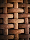 A close-up view of a textured woven pattern created by interlacing strands that resemble wood or plastic with a grainy texture. The strands are arranged in a horizontal and vertical crisscross fashion.