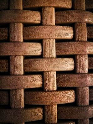 A close-up view of a textured woven pattern created by interlacing strands that resemble wood or plastic with a grainy texture. The strands are arranged in a horizontal and vertical crisscross fashion.