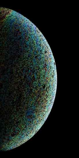 A large spherical object with a textured surface appears against a black background. The sphere displays a diverse array of colors, including blues, greens, and blacks, giving it a mysterious and cosmic appearance, reminiscent of a celestial body or planet.
