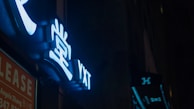 Neon signs with bright, glowing symbols and letters are mounted against a dark background. The signage prominently displays stylized characters in a blue hue, illuminating the surrounding area with their light.