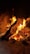A wooden log engulfed in bright orange and yellow flames, surrounded by glowing embers and ashes at the base. The setting is within a brick structure, suggesting a fireplace or kiln.