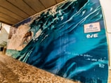 A vibrant mural depicting a surfer riding a large wave, with dynamic motion captured in the spray and curvature of the water. The mural is adorned with a sign that reads 'Praça dos Navegantes,' and includes artistic text stating 'O Surf Liberta as ondas... de uma vida!'. The scene conveys a sense of freedom and connection with nature.