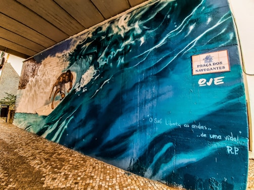 A vibrant mural depicting a surfer riding a large wave, with dynamic motion captured in the spray and curvature of the water. The mural is adorned with a sign that reads 'Praça dos Navegantes,' and includes artistic text stating 'O Surf Liberta as ondas... de uma vida!'. The scene conveys a sense of freedom and connection with nature.