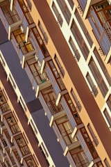 A high-rise building featuring a repetitive pattern of windows and balconies with wooden accents. The structure has a modern architectural design with a warm color palette and appears to be taken from an angled perspective.