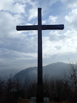 A quiet, humble cross standing in a simple rural landscape at dawn.