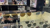 A display case with multiple pairs of sunglasses, including both aviator and classic styles. The sunglasses have varying lens colors and are arranged neatly for retail. In the background, the store environment is visible with shelves and a person walking by.