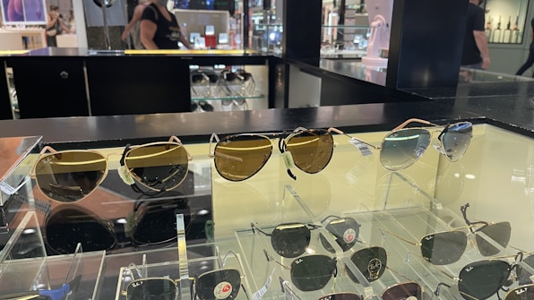 A display case with multiple pairs of sunglasses, including both aviator and classic styles. The sunglasses have varying lens colors and are arranged neatly for retail. In the background, the store environment is visible with shelves and a person walking by.