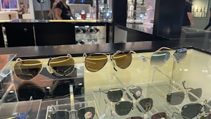 A display case with multiple pairs of sunglasses, including both aviator and classic styles. The sunglasses have varying lens colors and are arranged neatly for retail. In the background, the store environment is visible with shelves and a person walking by.